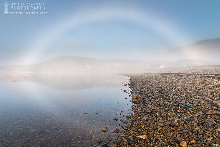 Fogbow, over pebble beach, sea loch and bothy reflected in calm water, Isle of Jura