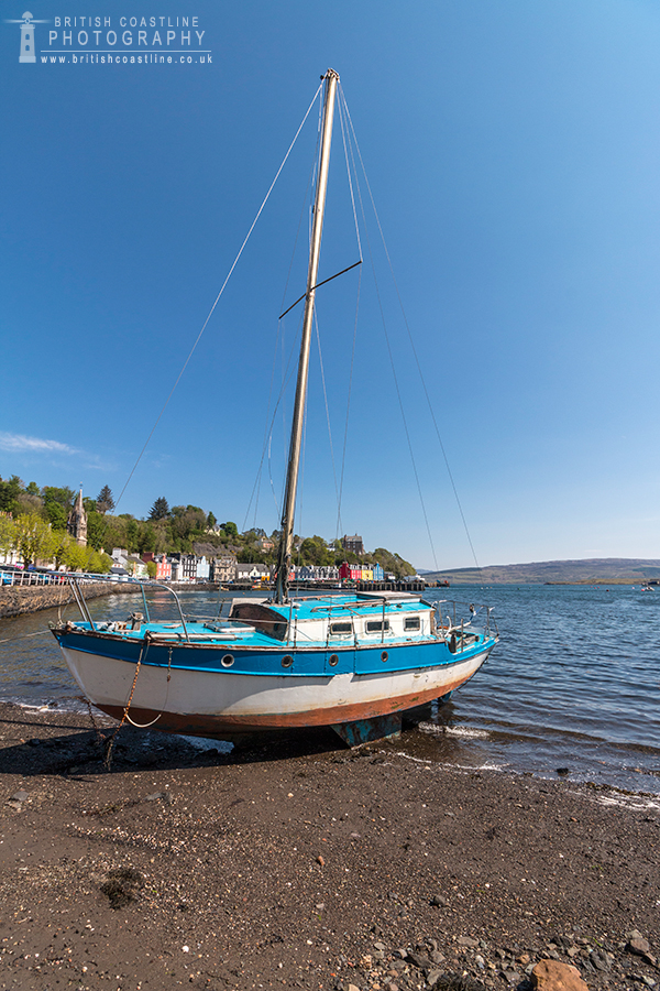 Tobermory, Isle of Mull, blue and white boat with tall mast grounded on beach with small waves lapping in, colourful houses, blue sky