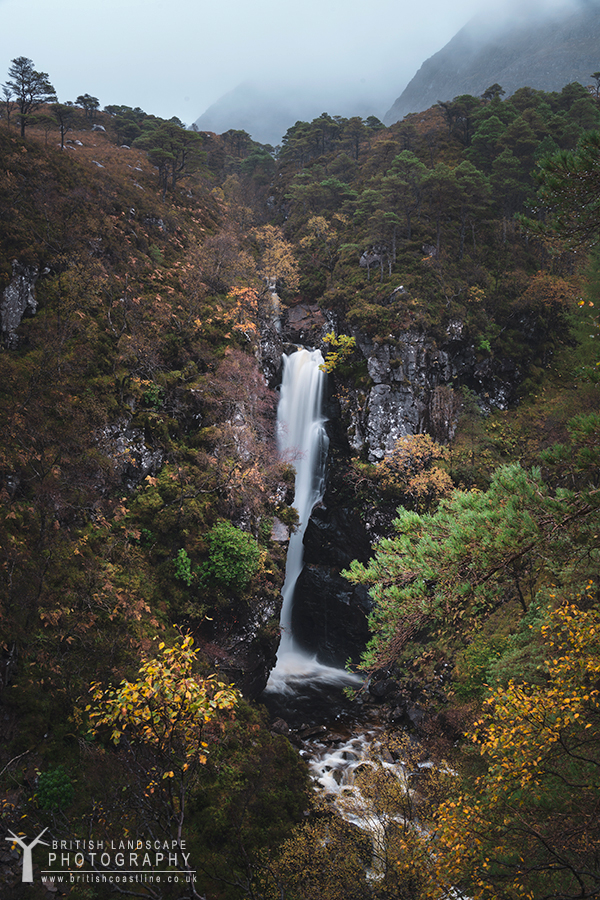 a long cascading waterfall with autumnal colours of surrounding forest and a mountaneous backdrop