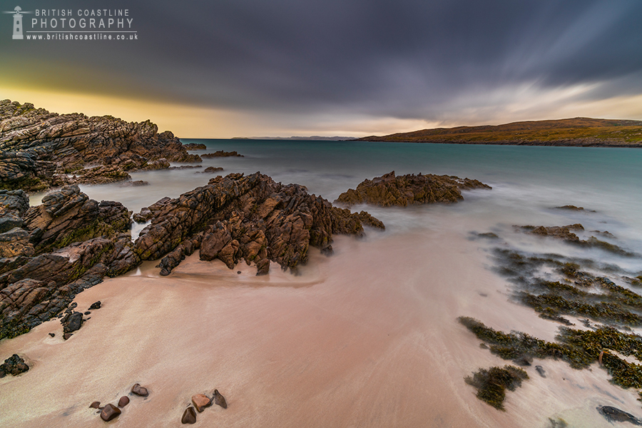 golden sandy beach, rocks draped with seaweed, blue turqoise sea, distant sea cliffs and clouds with motion