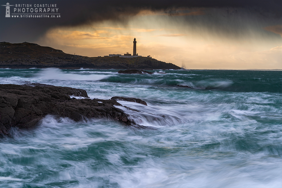 crashing waves on foreground rocks with a lighthouse on background rocks and storm like sky opening up to reveal golden light